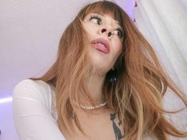 AriiannaHot with long brown hair, pearl necklace, and white top, tongue out, looking up with large eyelashes, showing her tits and tattoo on her chest.