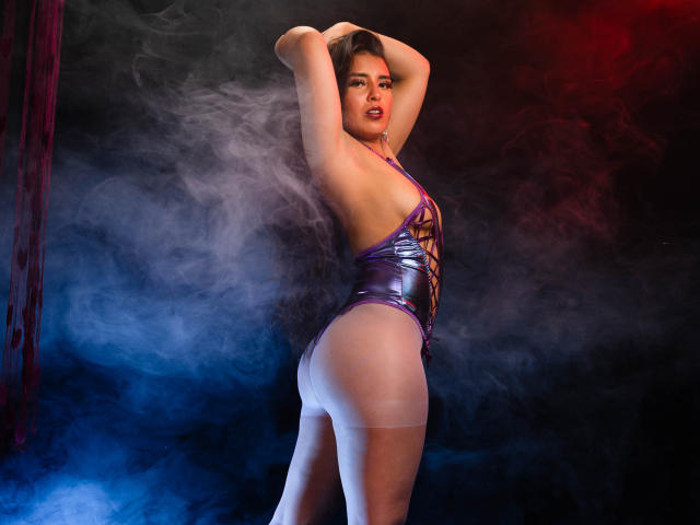 MelizaQueen indossa un costume intero viola scintillante, con le braccia alzate, mostra il sedere, è in piedi in un fumo blu e rosso e guarda seducentemente la fotocamera. Calze velate fino alla coscia, espressione sensuale..