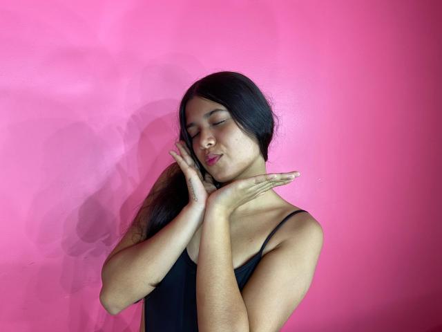 GenesisIsis, com os olhos fechados, cabelo preto e top preto, beijando os dedos contra uma parede rosa. Ela tem um nariz fofo e está fazendo um gesto de "silêncio"..
