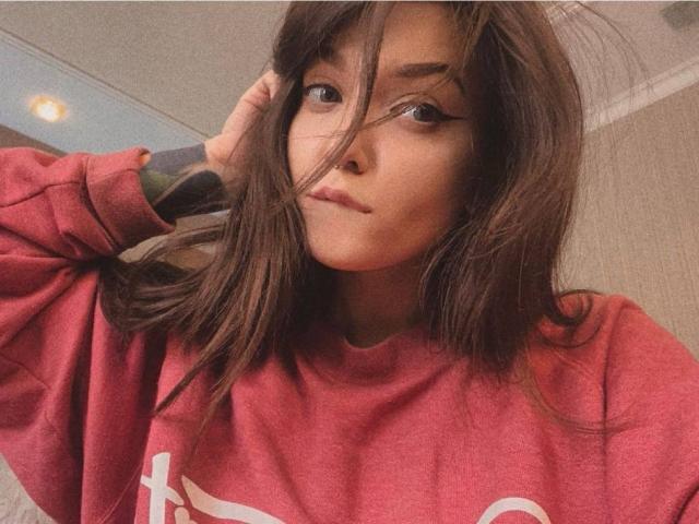 IrishReversed with medium-length brown hair, wearing a red sweatshirt, has a serious expression, and is touching her hair with her right hand. Her eyeliner is winged.