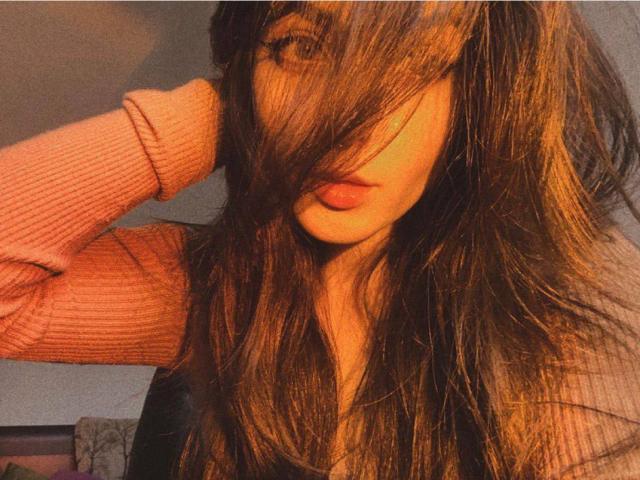 IrishReversed with long brown hair covering one eye, wearing a pink ribbed sweater, has her tongue slightly out, hand in hair, soft orange lighting.
