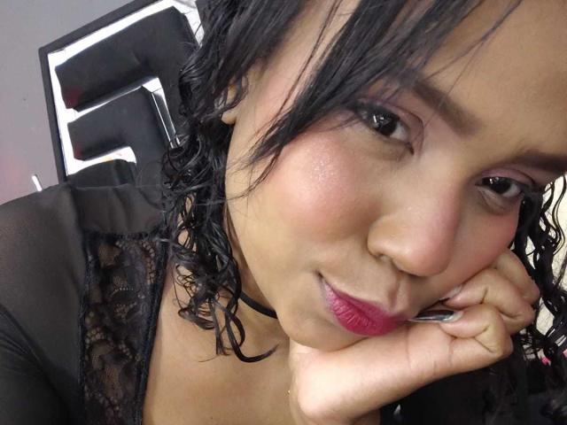 SucejPonce with curly black hair, pink lipstick, and a black lace top, resting her cheek on her hand, smiling at the camera, showing off her round ass.