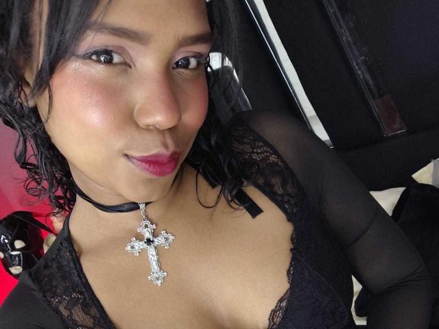 SucejPonce with medium brown skin, black lace top, pink lipstick, and black choker with diamond cross, showing small tits, curly black hair, and a slight smile.
