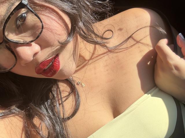 LunaSantos with red lipstick, glasses, and curly dark hair, showing off her boobs in a yellow strapless top, sunlight on her tan skin, and a necklace with small jewels.