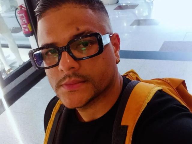 TheHottLover has a medium skin tone and short, dark hair. He wears thick, black, square-framed glasses and a small gold earring in his left ear. He has a trimmed mustache and goatee. He's dressed in a black shirt and a yellow backpack is slung over his shoulders. His expression is neutral. The image has a bright, reflective floor and a glass window in the background. Slightly blurry.