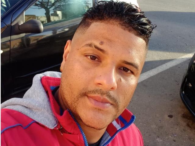 A close-up selfie of TheHottLover with medium brown skin, short curly black hair, and a small mole above the right eyebrow. He's wearing a red and gray zip-up hoodie. His expression is relaxed, with a slight smile. The sun casts a shadow on his face, highlighting his stubbled jawline. The image is sharp, with a clear focus on his face. The background includes a black car and a parking lot.