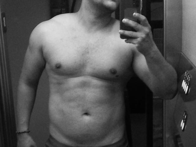 Il petto nudo e muscoloso di TheHottLover, con piccoli capezzoli e un leggero addome morbido, che tiene un telefono in un selfie allo specchio, indossa pantaloni scuri, in una foto in bianco e nero..