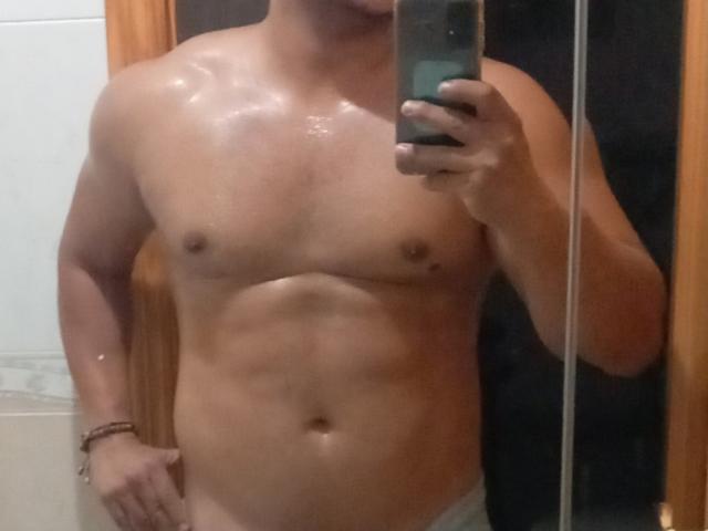 Shirtless selfie of TheHottLover, standing in front of a mirror. He has a muscular, defined chest and abs, with a slight sheen on his skin from sweat or oil. His right hand holds a green smartphone, partially covering his face. He wears only grey underwear, with a brown bracelet on his left wrist. The image focuses on his upper torso and lower abdomen. The background is blurred, emphasizing TheHottLover's physique. The mirror's reflection captures the details of his body.