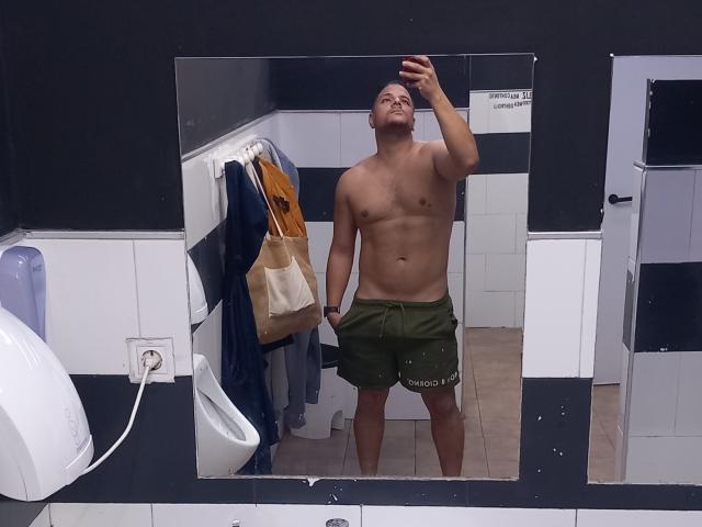 A mirror selfie of TheHottLover, a shirtless, muscular man with a fair skin tone and short dark hair. He's wearing green shorts with "Soccer" printed on them, standing with a confident, slightly tilted head. His right arm is raised, holding a phone to take the photo. A brown bag and blue shirt hang on a white wall-mounted hook behind him. The room has black and white tiled walls and a white urinal on the left. The image is clear, with sharp details and a casual, everyday setting.