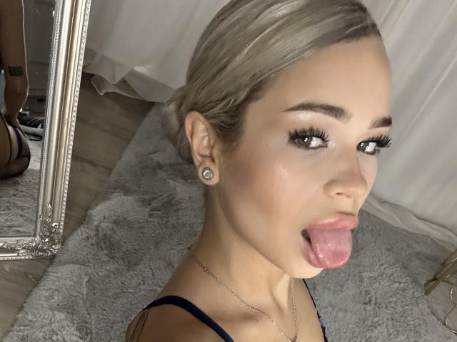 NickyBenz with blonde hair, green eyes, and earrings, sticking out her tongue, showing her pink tongue and slightly open mouth. She's wearing a blue top and a silver necklace.
