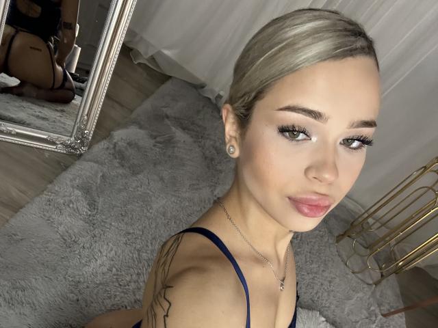 NickyBenz with blonde hair, blue eyes, and pink lipstick, wearing a blue top, showing tattoos, and a necklace, sitting on a gray rug, reflecting in a mirror.