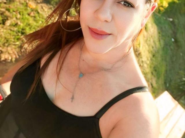 AlanaSunny with red lipstick, black tank top, and hoop earrings, showing her tits and a necklace with a blue gem and cross. Her brown hair is down.