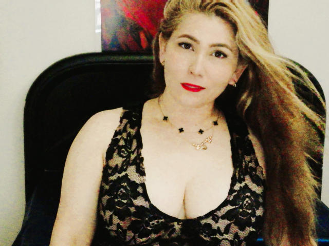 AishaSmithy with long blonde hair, red lipstick, black lace top, large boobs, star necklace, and gold chain, sitting in a black chair, looking at the camera.