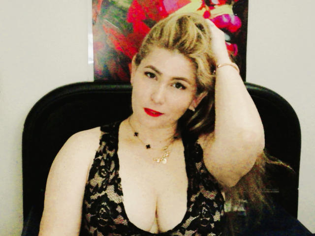 AishaSmithy with blonde hair, red lipstick, black lace dress, hand in hair, showing big tits, fair skin, black chair, gold necklace, and confident gaze.