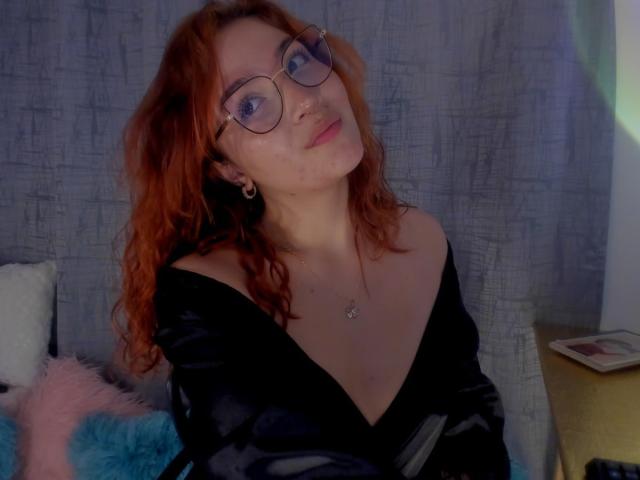 AmaruBloom with red curly hair, glasses, black off-shoulder shirt, and a necklace, looking up with a slight smile, showing her tits. Soft light, blue pillow, white pillow.
