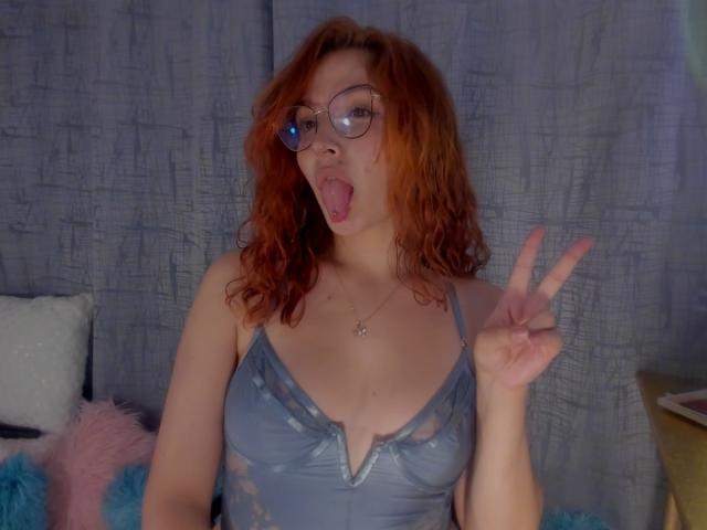AmaruBloom with red curly hair, glasses, and tongue out, wearing a grey lace dress, flashing a peace sign, showing her tits. Blue and pink pillows in background.