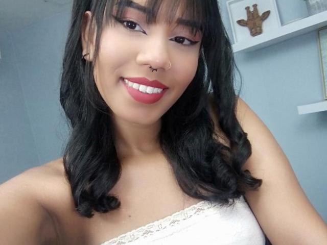 MiissDiiamondss has black wavy hair, brown eyes, red lipstick, nose ring, and smiling with white lace strap top, showing her tits. She's cute with a sexy look.