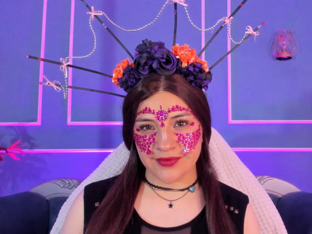 TaraTaylor has dark hair, purple and orange flower headpiece, pink glitter face makeup, black dress, white veil, black choker, sitting with purple neon lights in the background.
