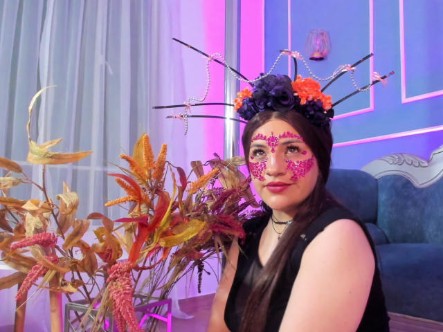 TaraTaylor has dark hair, pink glitter face paint, black sleeveless top, and a flower crown with orange and purple flowers, sitting next to autumn leaves, wearing a black choker.