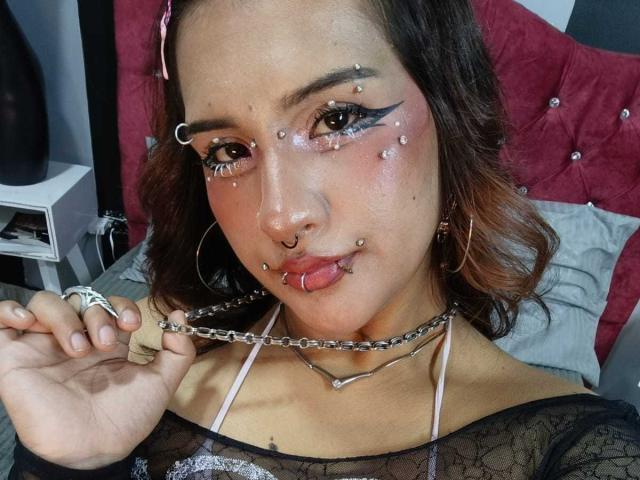 DanniLuke has piercings on her nose, eyebrows, and lip, with chain jewelry and dark makeup. She's holding a chain, wearing a black lace top, and sticking out her tongue.