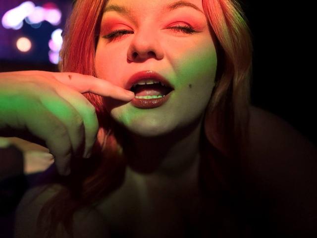 RomanMoore with red hair, eyes half-closed, lips slightly parted, finger touching her bottom lip, moles on her cheek, illuminated by green and red lights.