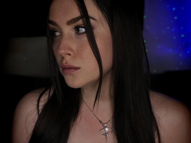 AliceOfWonderss has long, straight black hair partially covering her right eye. She has light skin with freckles, full eyebrows, and wears subtle makeup. Her lips are slightly parted, and she looks to the left. She's wearing a silver star necklace with a small white gem in the center. The dark background contrasts with a faint blue light on the right. Her expression is neutral, and the image is focused on her upper torso.