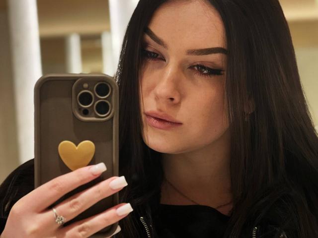 Close-up of AliceOfWonderss with straight, dark brown hair, focused on a smartphone. She has light skin, pronounced eyebrows, and subtle makeup with winged eyeliner and pink lipstick. She wears a black top, and a gold heart-shaped phone case is visible on the phone. Her nails are long and white, with a silver ring on her index finger. The image has a soft, warm light.