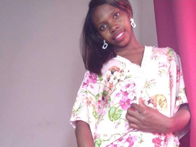 ElianeJolie with dark brown skin, wearing a pink floral shirt, white earrings, and smiling, touching her chest, standing against a pink wall. Her tits are partially visible.