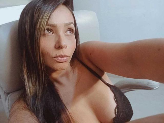 LuluSinclair with long black hair and brown highlights, wearing a black lace bra, has medium tits and a serious expression, sitting in a white chair, taking a selfie.