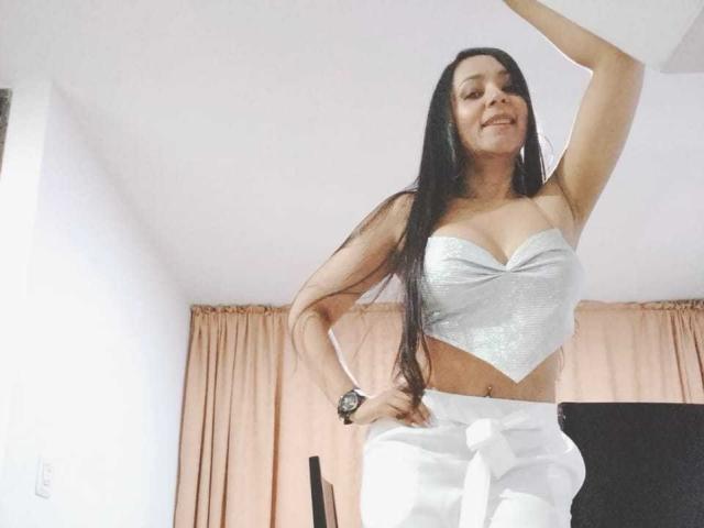 LuluSinclair, avec ses longs cheveux noirs, portant un soutien-gorge argenté brillant et un pantalon blanc, souriante, une main sur la hanche, levant le bras, une montre noire au poignet, peau claire et une jolie fesse..