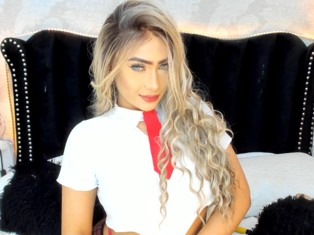 VictoriiaGrey with long, wavy blonde hair, red lipstick, white shirt, and red tie, sitting on a bed with a black, studded headboard. She's looking at the camera.