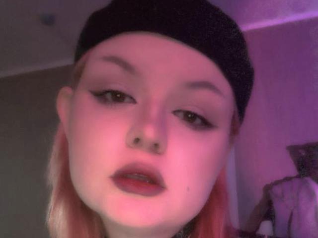 RomanMoore with pink hair, black beanie, and thick eyeliner, staring into the camera with slightly parted pink lips, pinkish-purple lighting on her pale skin.
