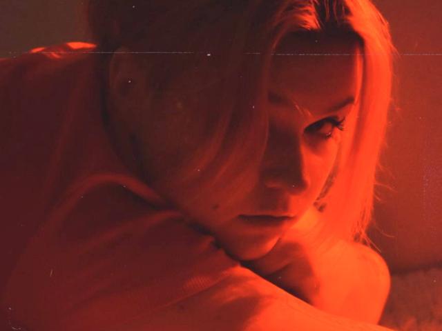 RomanMoore's face is bathed in red-orange light, lying down with eyes partially closed, looking seductive. Her blonde hair is slightly messy, and she's wearing a red shirt.