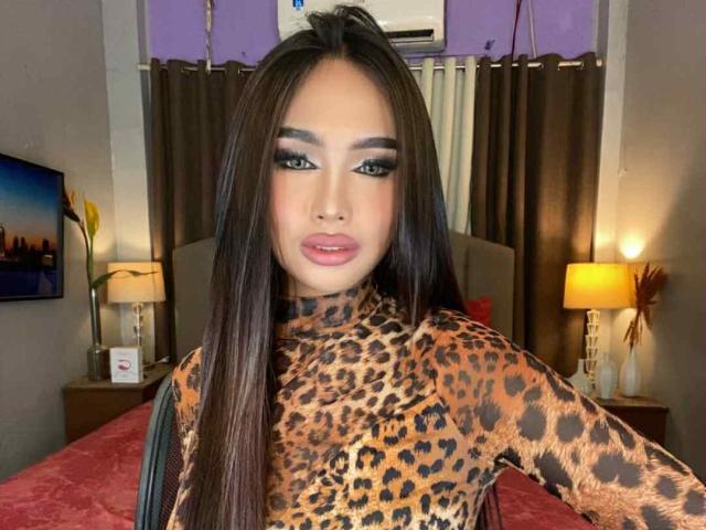 BigBrownCockCandice with long brown hair, wearing a leopard print top, showing off her makeup with bold eyeliner and pink lipstick, standing in a bedroom with lamps.