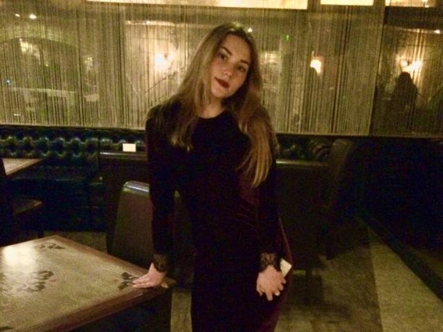 CaseyMeli with long brown hair, wearing a black dress, leaning on a table, hands on her ass, slightly tilted, in a dimly lit bar.