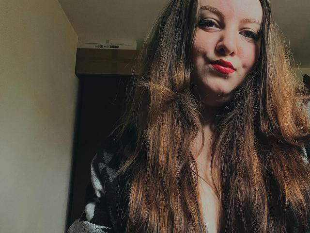 CassiaMoon with long brown hair, red lipstick, and a subtle smile, wearing a gray and black striped shirt, looking down at the camera.
