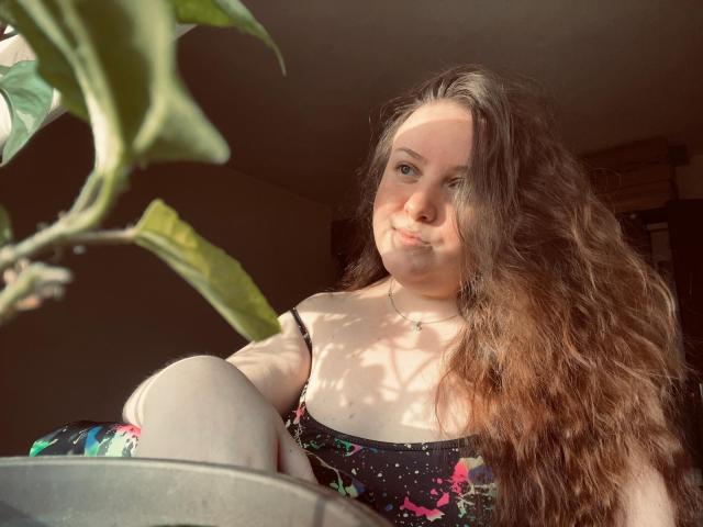 CassiaMoon with long, wavy brown hair, fair skin, and small tits, wearing a floral black dress, sitting by a plant, sunlight on her face, smiling gently.