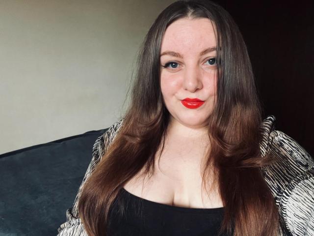 CassiaMoon with long brown hair, red lipstick, blue eyes, and cleavage in a black top, wearing a striped jacket, smiling at the camera.