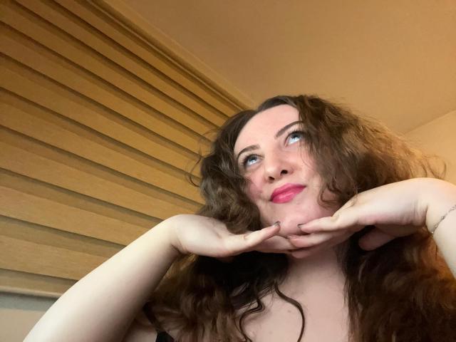 CassiaMoon with wavy brown hair, pink lipstick, and raised hands, looking up with a soft smile. She's got a cute ass, and her tits are subtle in a black top.