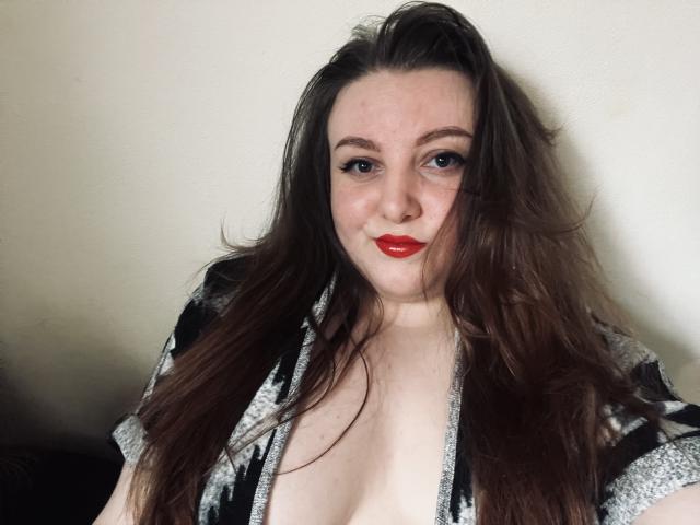 CassiaMoon with long brown hair, red lipstick, and black/white patterned top, showing her tits, standing against a plain wall, looking at the camera with a slight smile.