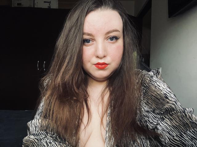 CassiaMoon has red lips, blue eyes, and long brown hair, wearing a zipped-up black and white striped top, showing a little cleavage. Her face is slightly flushed.