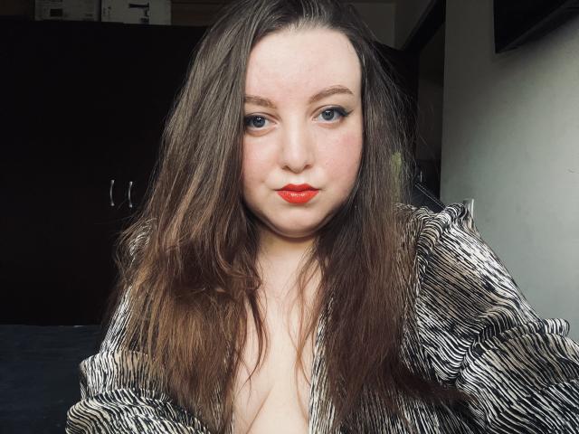 CassiaMoon has long brown hair, red lipstick, and blue eyes, wearing a striped shirt, with her tits slightly visible, in a selfie.