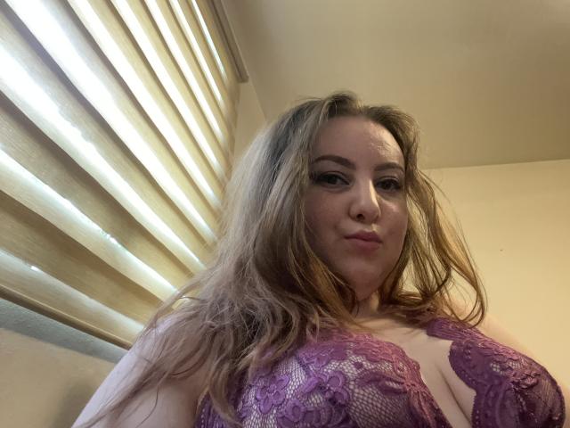 CassiaMoon with long blonde hair, wearing a purple lace bra, showing her big tits, looking down at the camera with a slight smile. Light from blinds on the left.