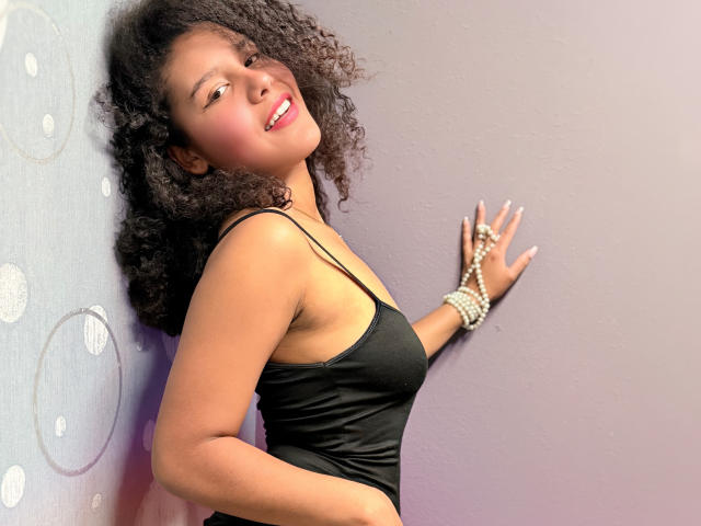 AriaVega, a young woman with curly black hair, stands against a purple wall, smiling. She wears a black spaghetti-strap tank top, showing a slender build and medium-sized breasts. Her right hand, adorned with a pearl bracelet and rings, touches the wall. The light highlights her warm brown skin. Her red lipstick contrasts with her natural look. The image has a soft, warm light, emphasizing her relaxed, confident pose.