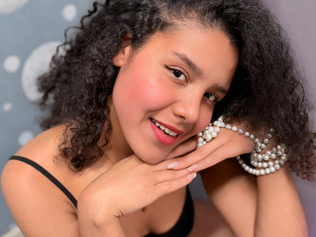 AriaVega with curly black hair, brown skin, and a cute smile, wearing a black bra and pearl bracelets, resting her chin on her hand, showing her tits slightly.