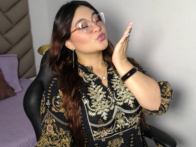 HelueJamil with glasses, dark wavy hair, and pink lipstick, touching her cheek with a manicured hand, wearing a black floral dress, sitting in a chair. Boobs visible.