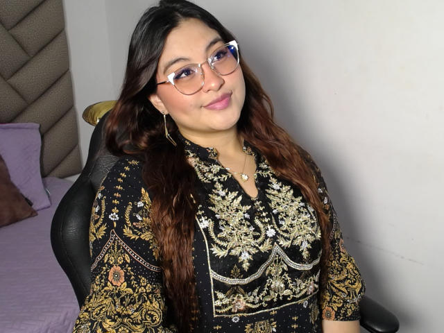 HelueJamil has brown wavy hair, glasses, and a black dress with gold embroidery, showing her tits, with a small necklace, and a subtle smile in a bedroom setting.
