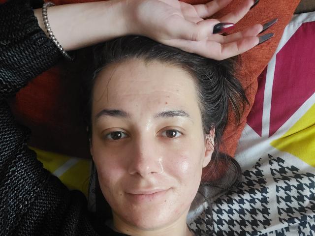 Julia Hunt, avec son visage brillant de sueur, son vernis à ongles noir, ses cheveux foncés, ses yeux marron, son pull noir, une main avec une bague, allongée sur un lit coloré, avec un sourire détendu..