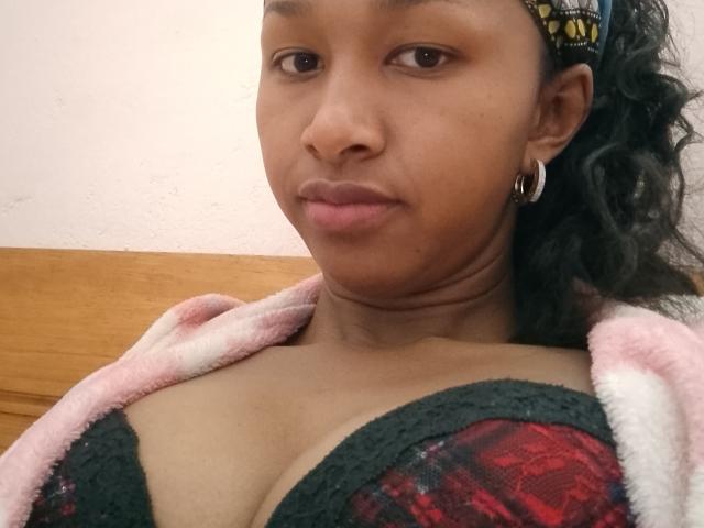 HotLorie with dark skin, curly green hair, wearing a green lace bra with red plaid pattern, pink and white fleece, and gold hoop earrings, looking at camera.