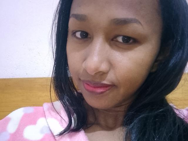 HotLorie has dark brown skin, black straight hair, and red lipstick. She's wearing a pink and white patterned shirt, with a wooden headrest behind her.
